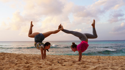 Two young girls gymnasts perform a gymnastic element on the beach of the Pacific Ocean. Flexible sports models in bright clothes at sunset perform a handstand. Seascape girls on the seashor