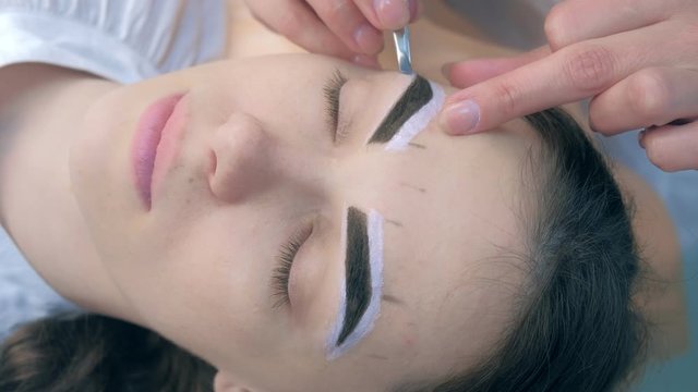 Professional beautician plucking eyebrows with tweezers to woman in beauty salon during tint eyebrow procedure, face closeup. Girl lying with closed eyes and brown natural henna on brows.