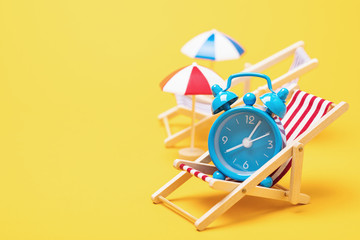 Alarm clock in a deck chair with a parasol on a yellow background with copy space. Concept on the...
