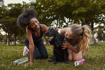 Two smiling young diverse female athletic friends playing and loving a black dog in the park 