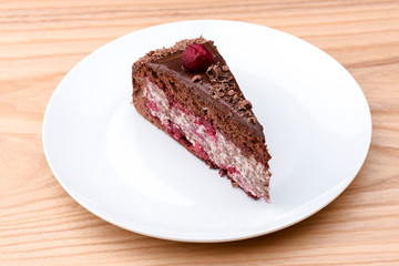 One piece of chocolate cake with vanilla cream and cherry topping served on white plate over light wooden background.