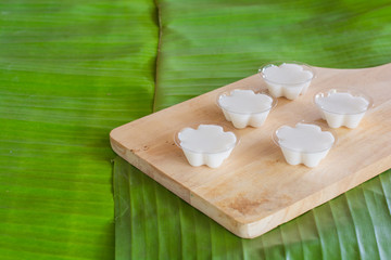 Colorful of Thai coconut jelly in small plastic cup on wooden plate with green banana leave background. Thai dessert.