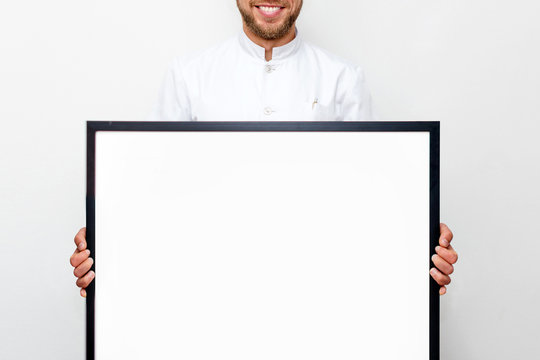 Man in white uniform holding a picture frame or poster for mock up. Doctor or chef smiling and showing empty frame, business and advertisement concept - man with white blank board