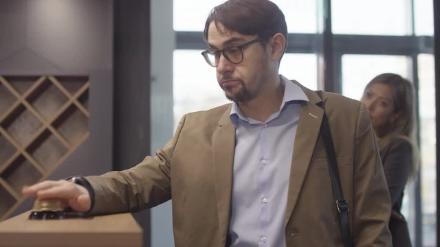 Tracking with slowmo of annoyed man in glasses standing at hotel reception desk and ringing bell, then looking at his wristwatch and sighing. Frustrated people queuing behind him
