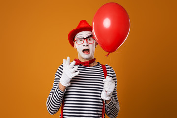 Clown mime in vest and red hat with balloon in his hand on orange background