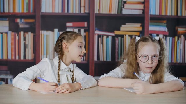 Schoolgirl copying solution from classmate at exam in school on bookcase library background. Tricky student girl peeking test answer in cheat sheet in classroom