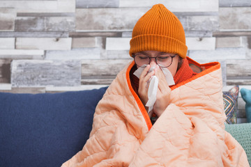 A sick teenager girl with a runny nose, sits on the couch and blows her nose. At home.