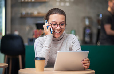 Cheerful teen girl talking on a cell phone at a table in a cafe. During a conversation, she laughs and looks at the tablet. In the background is the interior of a cafe.