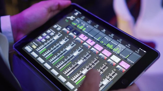 DJ sound control in application on the tablet for mixing instruments of music band in disco club concert hall. Hands touching buttons sliders, levels of volume changing. Color illumination
