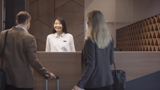 Slowmo tracking shot of man and woman with suitcases walking up to reception desk at hotel and talking to cheerful Asian female receptionist