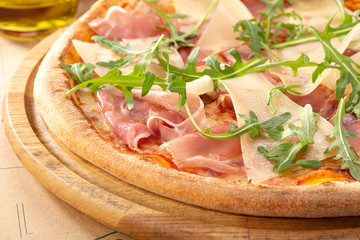 Delicious fresh pizza on table. Pizza with prosciutto (parma ham), arugula (salad rocket) and parmesan, served on a wooden table for dinner, close up. Italian cuisine.