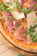 Delicious fresh pizza on table. Pizza with prosciutto (parma ham), arugula (salad rocket) and parmesan, served on a wooden table for dinner, close up. Italian cuisine.