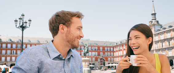 Couple drinking coffee at Madrid cafe dating laughing panoramic banner. Two tourists peaple having fun conversation talking on outdoor Plaza Mayor terrace summer travel vacation. Young woman and man.