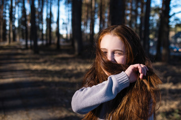 Little cute girl with long red hair posing in a pine park.
