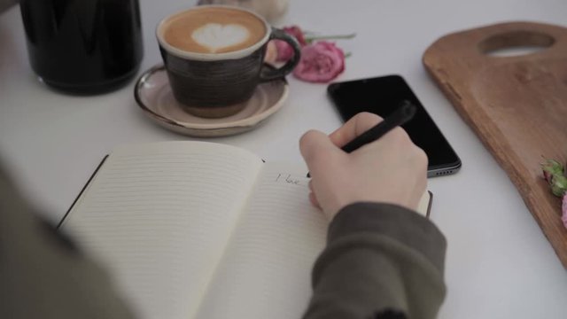Girl's hand writes I love you in a notebook that is on a white table next to a cup of coffee and a phone