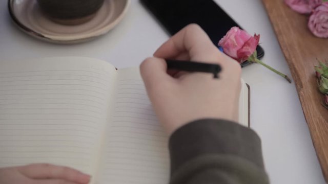 Girl's hand writes I love you in a notebook that is on a white table next to a cup of coffee and a phone