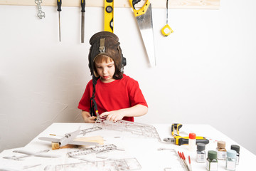 Children's hobbies: A little boy in a pilot's helmet builds a large-scale model of an airplane from plastic parts using a circuit. - Powered by Adobe