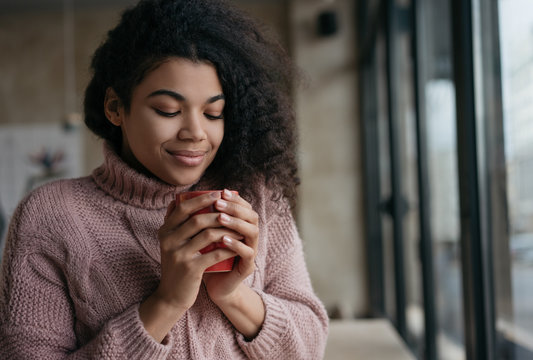 Portrait of young beautiful African American woman holding cup of coffee near face. Pensive hipster girl with eyes closed drinking tea, sitting in cafe