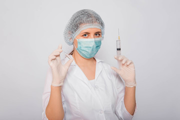 The doctor is holding a syringe and an ampule with a vaccine. Young woman in a white coat, in a medical mask, hat and gloves on a white background. Laboratory medical worker.