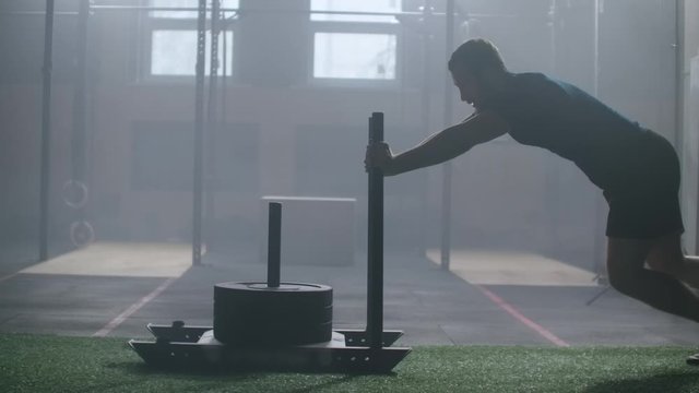 Young strong athletic Caucasian man exercising with heavy weight training sled towards camera in large gym slow motion