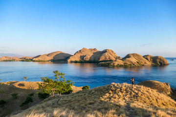 Fototapeta na wymiar A woman standing on top of a small island, enjoying the morning sun over Komodo National Park, Flores, Indonesia. Golden hour over the islands and sea. Some boats anchored to the bay. New day 