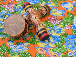 Three colorful handmade Brazilian percussion instruments: a small drum and two "ganzás", a type of rattle, on a colorful “chitao” fabric with floral prints. They are widely used in Carnival.