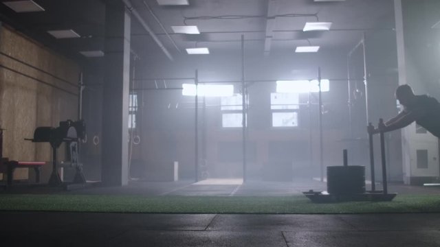 Willpower and motivation, camera follows young athletic man pushing heavy training sled training in gym slow motion