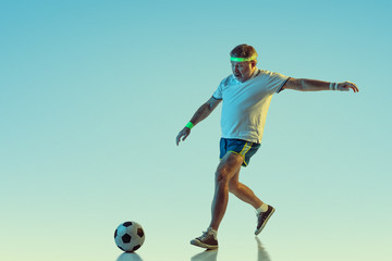 Senior man playing soccer, football on gradient background in neon light. Caucasian male model in great shape stays active, sportive. Concept of sport, activity, movement, wellbeing, healthy lifestyle
