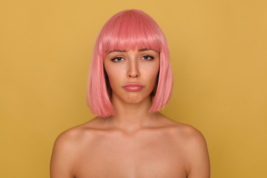 Wailful young attractive pink haired female with short haircut looking sadly at camera and pouting her lips while standing over mustard background with naked shoulders