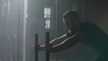 Willpower and motivation, camera follows young athletic man pushing heavy training sled training in gym slow motion