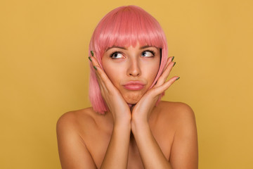 Indoor shot of young pretty blue-eyed lady with pink bob hairstyle holding her face with raised palms and looking upwards with puzzled face, posing over mustard background