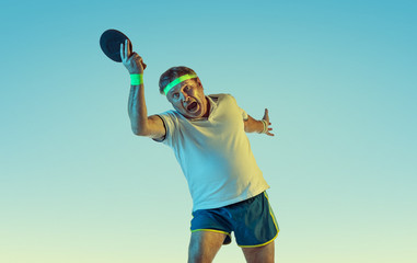 Senior man playing table tennis on gradient background in neon light. Caucasian male model in great shape stays active, sportive. Concept of sport, activity, movement, wellbeing, healthy lifestyle.
