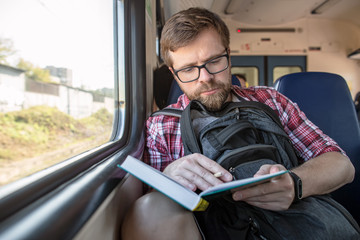 Focused bearded man riding in a train, reading a book and making notes with a pencil.