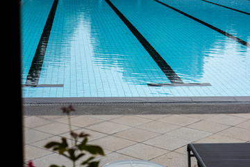 A swimming pool with a sunny reflection and beautiful wavy water surface in turquoise blue colour tiles and black line lanes underwater. Nice tree reflection on the pool surface.