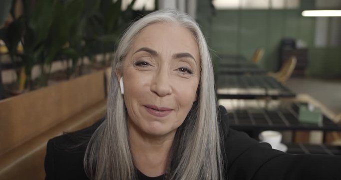 Close up of elegant middle aged businesswoman in earpods having videocall on smartphone. Camera view of beautiful old woman communicating, using video app of her phone while sitting in cafe.
