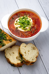 Russian red borscht with sour cream and greens in white bowl and pieces of bread on wooden background, table. Ukrainian cuisine concept.