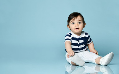 Baby boy in striped t-shirt, white pants and booties. He is smiling, sitting on floor against blue studio background. Close up