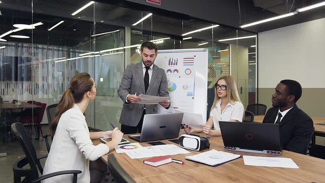 Attractive experienced bearded businessman in suit standing near flip chart presentation and talking to his business multiethnic industrious team