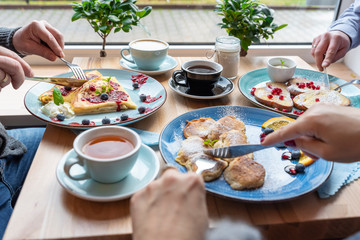Delicious colorful meals for breakfast in the morning with hands of people and cutlery. Plates of healthy food and cup of coffee and tea. Meeting during the lunch time. Close up. 