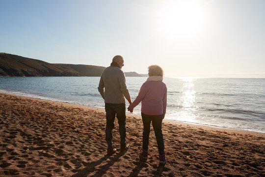 Rear View Of Loving Retired Couple Holding Hands Looking Out To Sea On Winter Beach Vacation