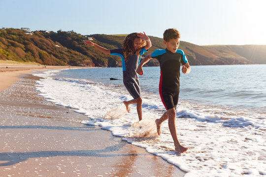 Two Children Wearing Wetsuits Running Through Waves On Summer Beach Vacation