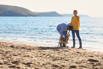 Couple Walking With Dog Along Shoreline On Winter Beach Vacation
