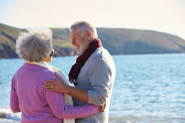 Loving Retired Couple Hugging Standing By Shore On Winter Beach Vacation Against Flaring Sun