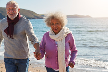 Active Senior Couple Running Along Winter Beach By Waves Holding Hands