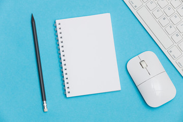 A blank white notebook for notes with a pencil on a blue background surrounded by a keyboard, headphones and mouse. Creative flat lay photo of workspace desk with copy space. 