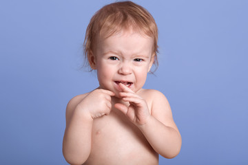 Horizontal shot of beautiful baby crying isolated over blue background, posing bared and crying, looking at camera, finding her mother, wants falling asleep and drinking milk, keeps finger in mouth.