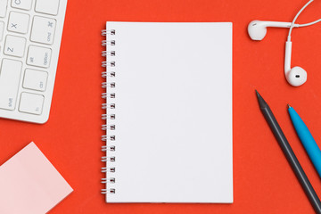Blank notebook on a red desktop surrounded by a keyboard, mouse, headphones and pencil. Creative flat lay, mock up