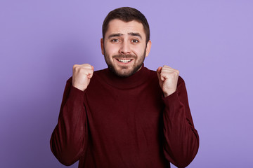 Fototapeta premium Indoor portrait of emotional charismatic good looking man with beard clenching fists, making gesture, smiling sincerely, having fun, getting good news, being in high spirits. People concept.