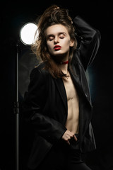 Beautiful braless girl with red lips, wearing an unbuttoned black blazer, touches her hair with her hand on a dark background, posing next to a light lamp. Advertising, trendy, and lifestyle design.