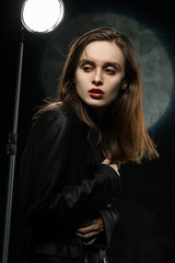 Fototapeta na wymiar Beautiful slim girl model with red lips, wearing a black blazer, fearfully hugs herself with hands on a dark background, posing next to the light lamp. Advertising, trendy, and lifestyle design.